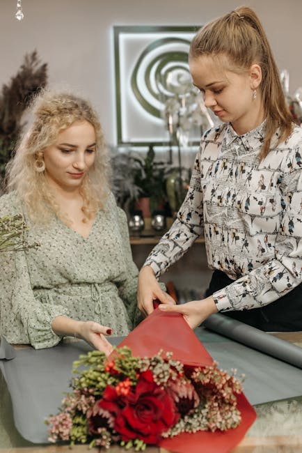 Two women are engaged in creating a floral gift, with one woman wrapping a bouquet of vibrant red roses, pink and white flowers, and green foliage in red wrapping paper. The arrangement features fresh