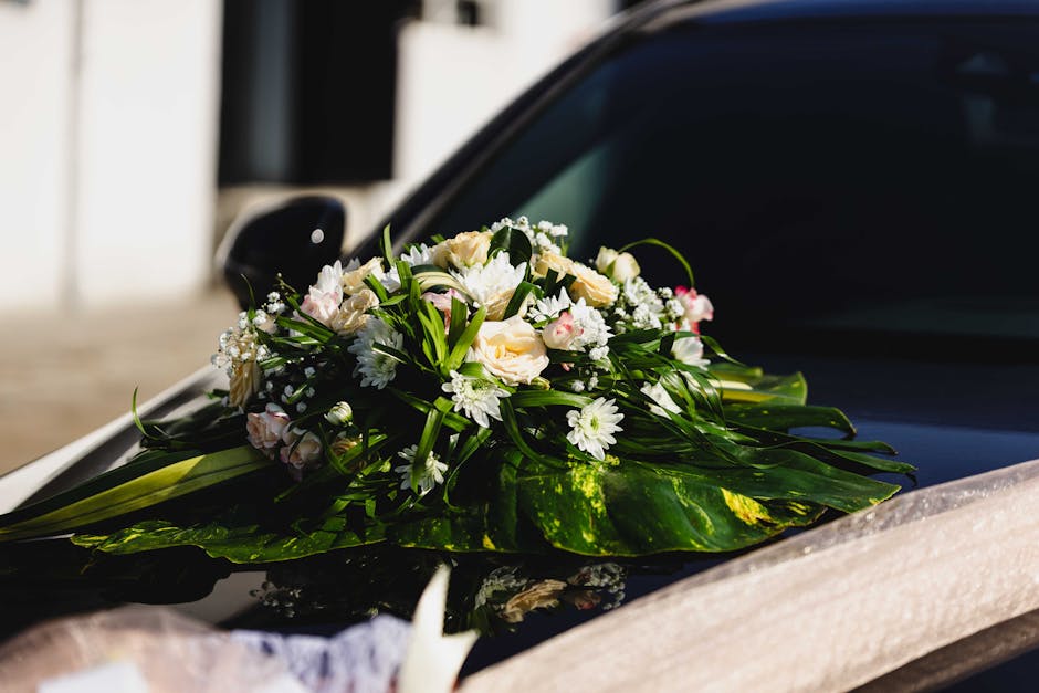 A fresh floral arrangement placed on the hood of a black car, featuring a bouquet of white and pale pink roses, white daisies, and delicate baby's breath, accented with lush green leaves and long gras