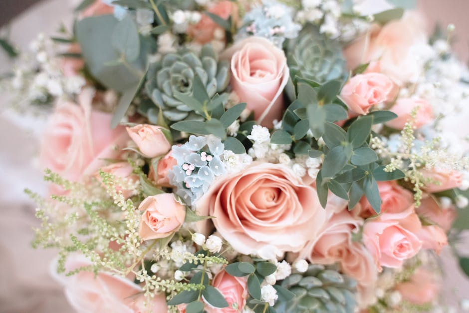 A close-up photograph of a floral arrangement featuring soft pink roses with tightly spiraled petals, complemented by delicate white baby's breath and pale blue sedum succulents. The bouquet is adorne