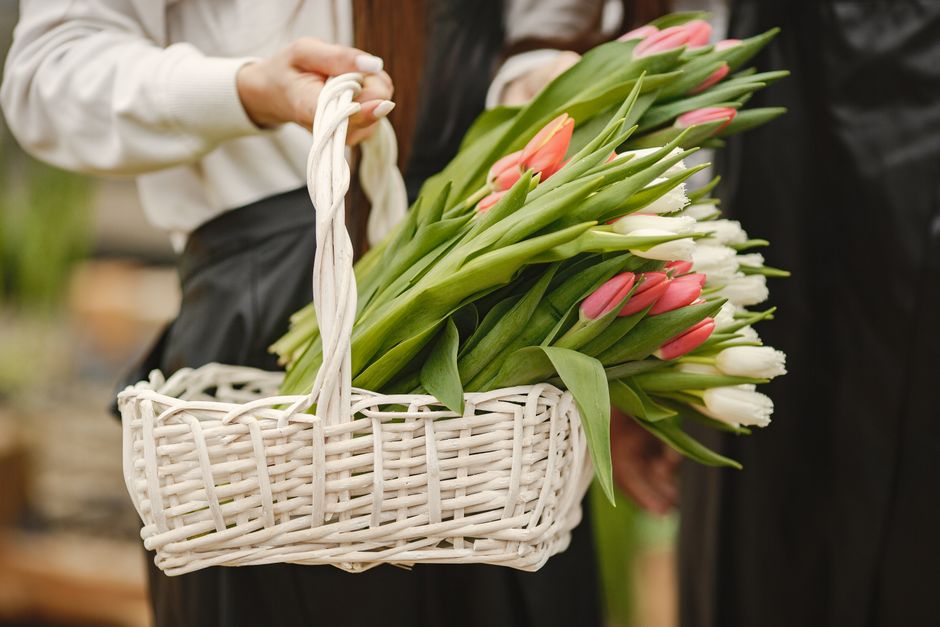 A person holding a white woven basket filled with fresh tulips in shades of pink and white, with green stems and leaves visible. The basket is being held by a hand wearing a cream-colored long-sleeve