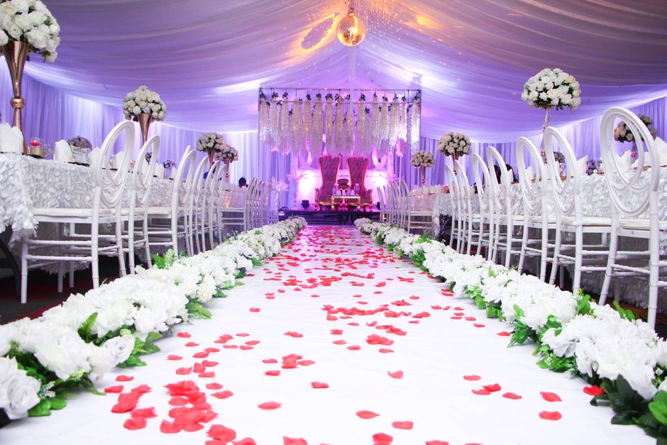 A luxurious wedding ceremony setting at Painshill Park featuring a central aisle covered with a white runner scattered with pink rose petals. Flanking the aisle are large, lush arrangements of white r