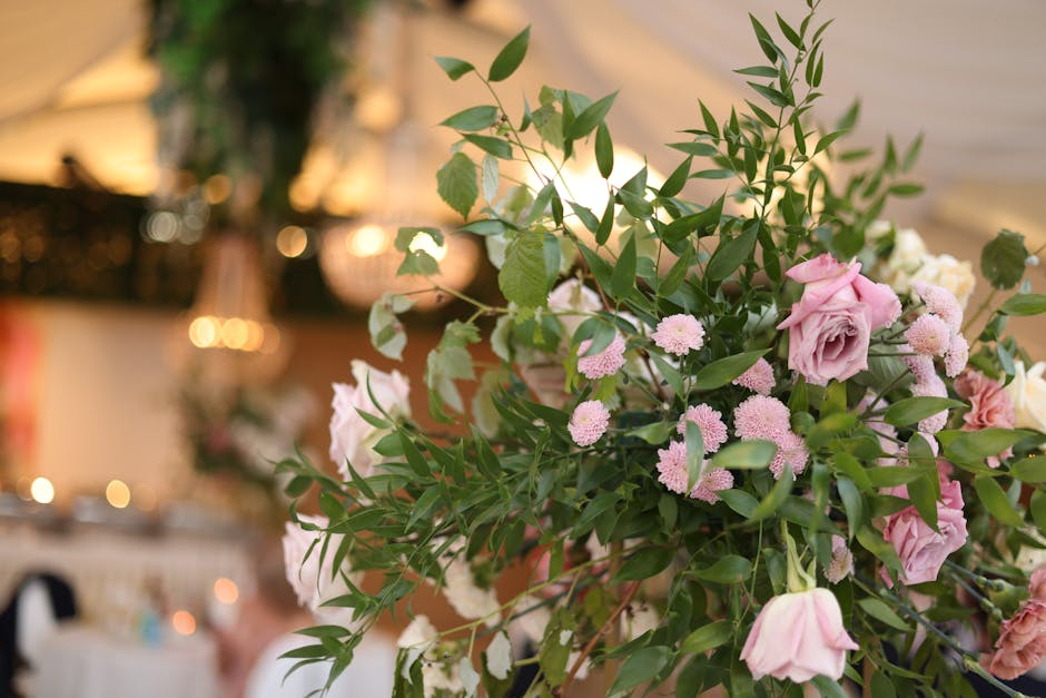 A close-up of a floral arrangement featuring soft pink roses and small pink pom-pom chrysanthemums intertwined with lush green foliage, arranged in a natural, cascading style. The bouquet is placed in